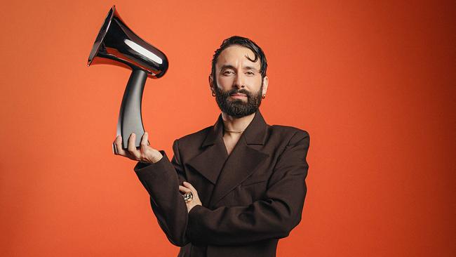 Tom Neuwirth in einem dunklen Anzug posiert vor orangefarbenem Hintergrund und hält die "Amadeus Austrian Music Awards 2026"-Trophäe in einer Hand.