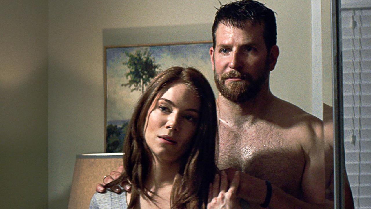 Eine Frau und ein Mann mit nacktem Oberkörper stehen nebeinander und blicken in einen Wandspiegel, hinter ihnen ist ein Gemälde und eine Lampe zu sehen