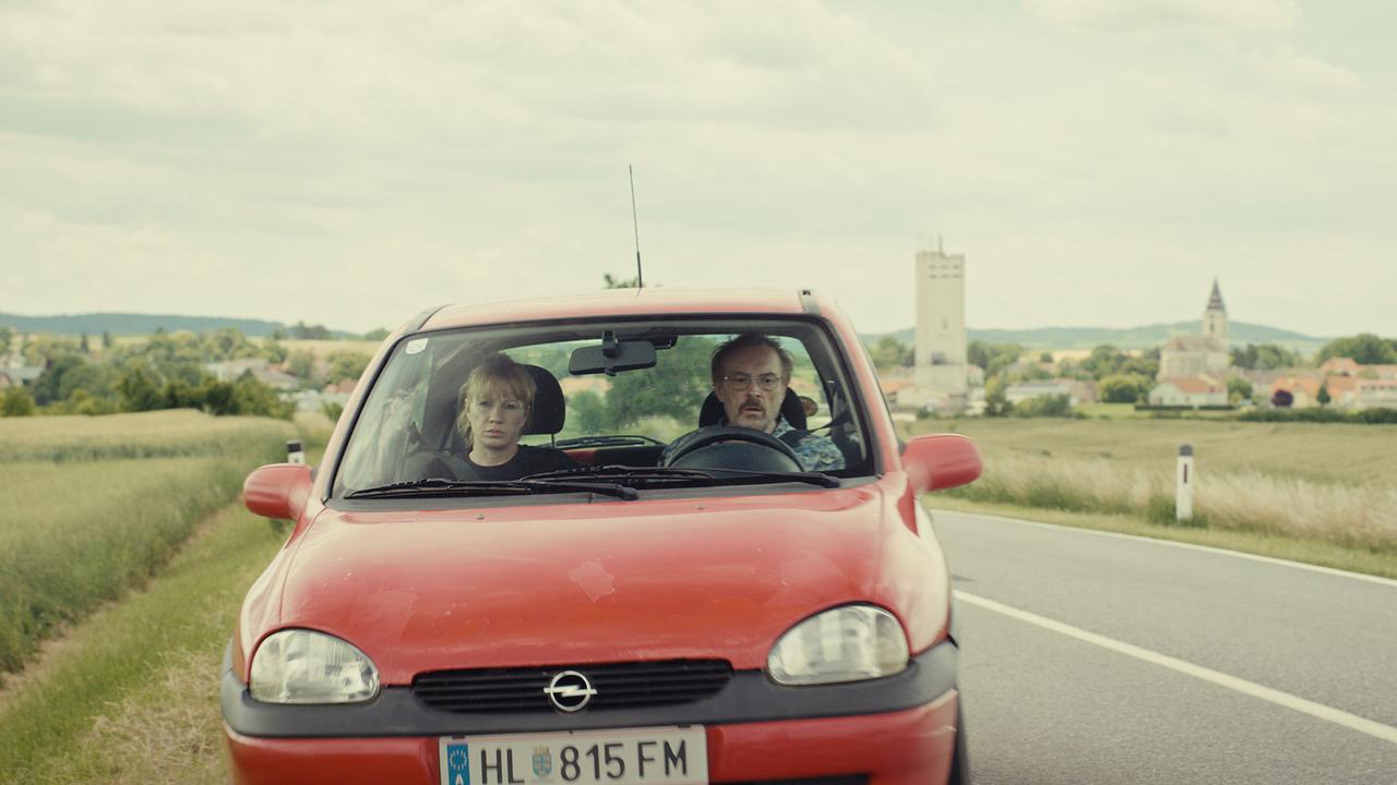Andrea und Franz fahren tagsüber in einem roten älteren Opel auf einer Landstraße, dahinter ist ein Ort mit Kirchturm zu sehen.
