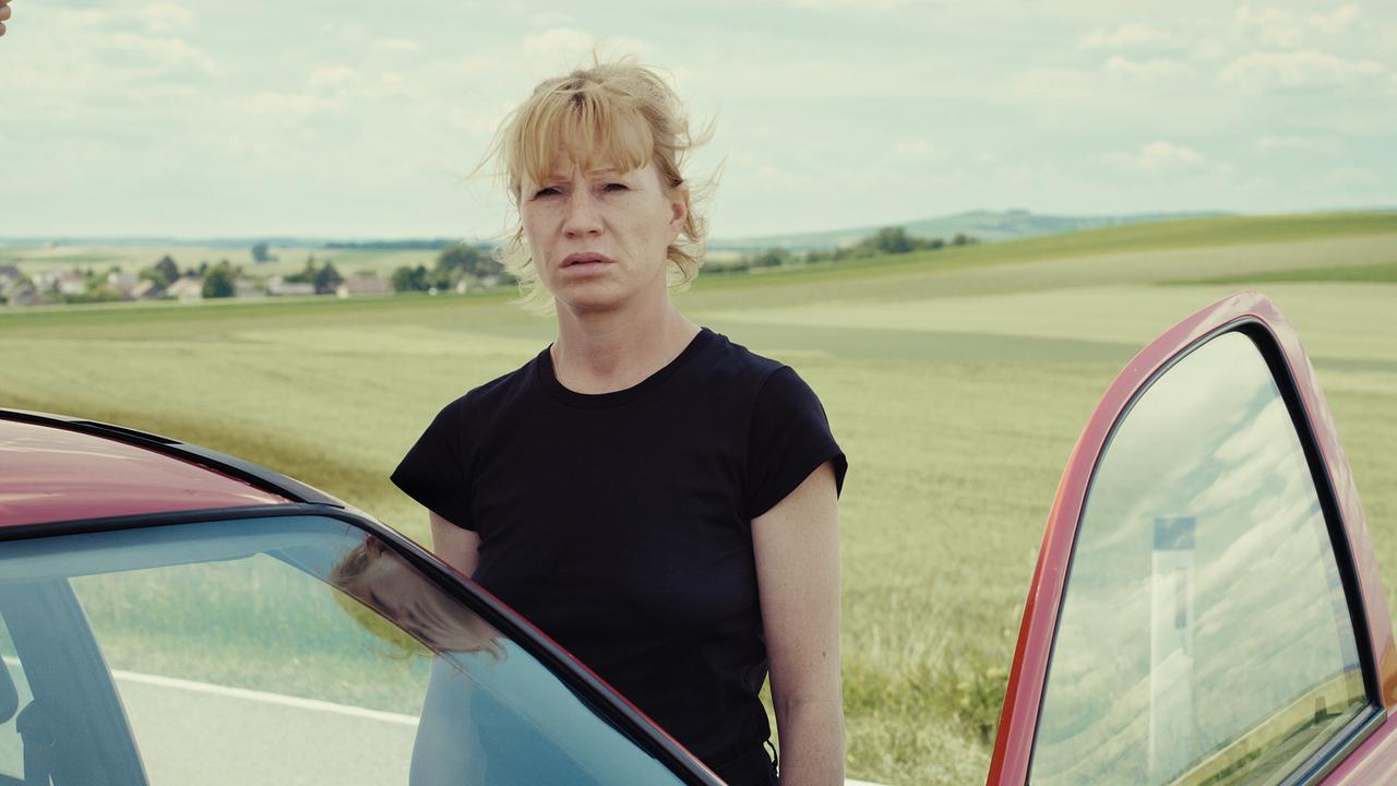 Andrea steht neben einem roten Opel mit geöffneter Tür vor einer weiten Hügellandschaft.