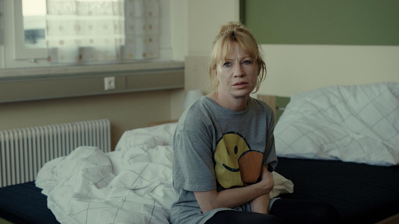 Andrea sitzt am Tag auf einem Bett in einem schlichten Zimmer und hält die Arme eng an den Körper.