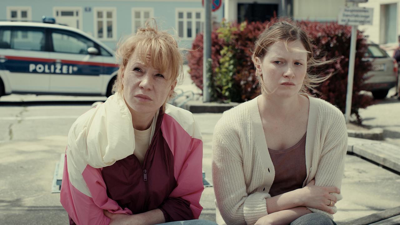 Andrea und Gerti sitzen draußen vor einem Gebäude auf einem Platz, im Hintergrund steht ein Polizeiauto.