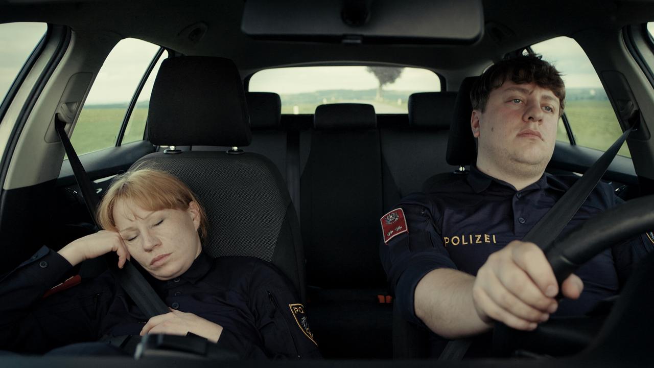 Polizistin Andrea sitzt dösend neben ihrem Kollegen Georg am Beifahrersitz und sie fahren über eine Landstraße, im Hintergrund ist eine dunkle Rauchwolke zu sehen.