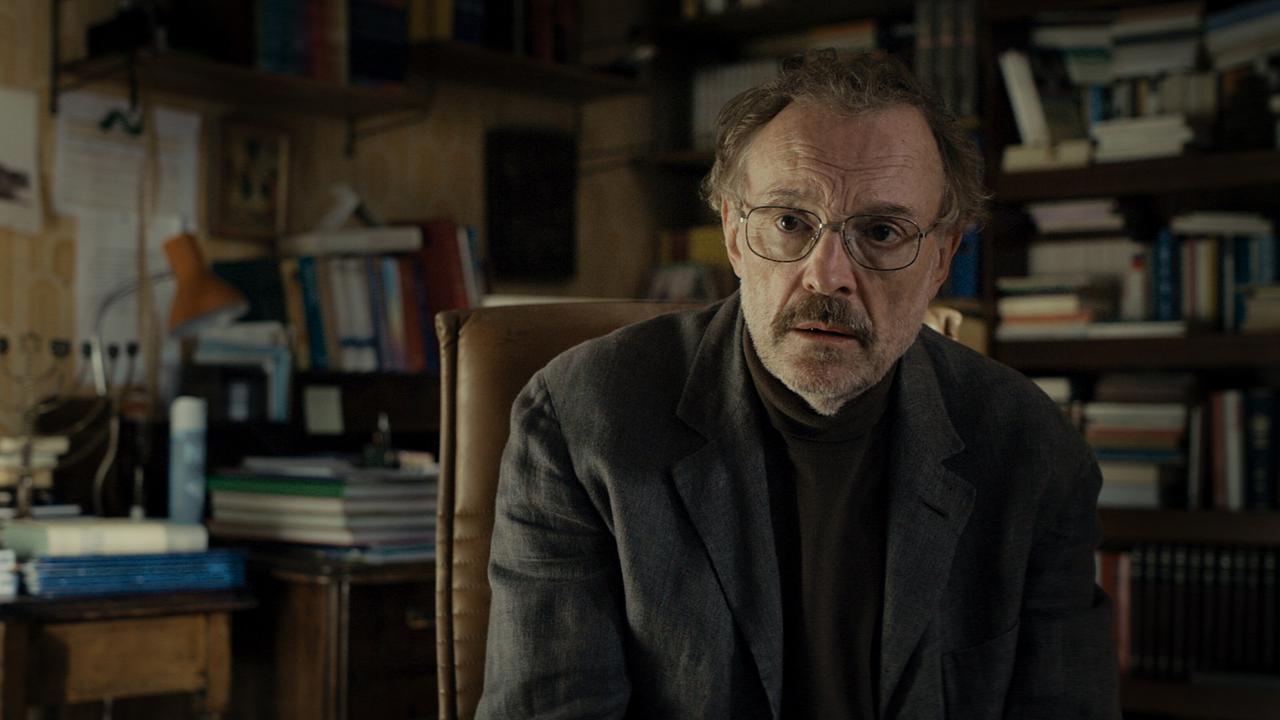 Franz sitzt in einem vollgestellten Arbeitszimmer auf einem Lederstuhl, umgeben von Büchern und Papieren.