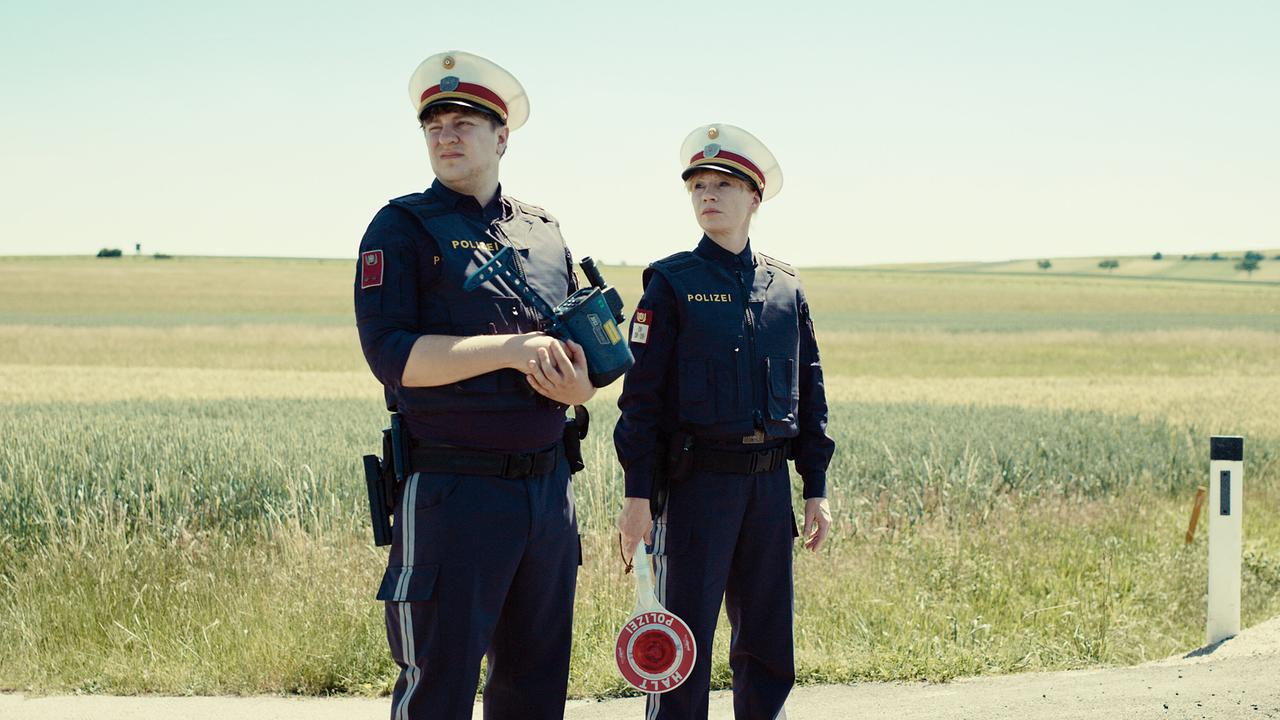 Georg und Andrea, beide in Polizeiuniform, stehen am Straßenrand neben Feldern, Georg hält eine Radarpistole und Andrea eine Anhaltekelle.