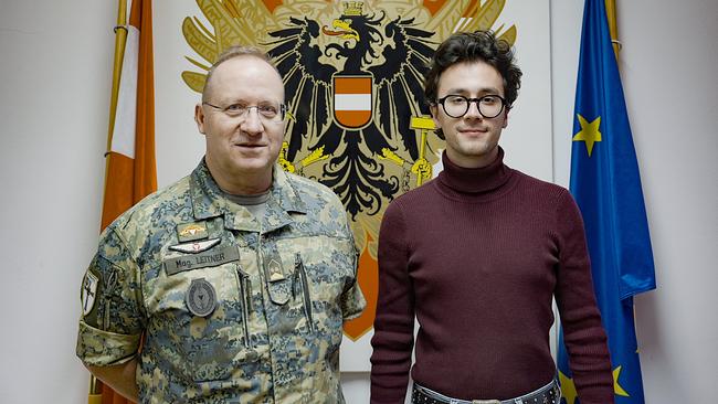 Lukas Leitner und Rocco Bald stehen vor einem großen Emblem der österreichischen Garde, flankiert von einer Österreich‑ und einer EU‑Fahne.