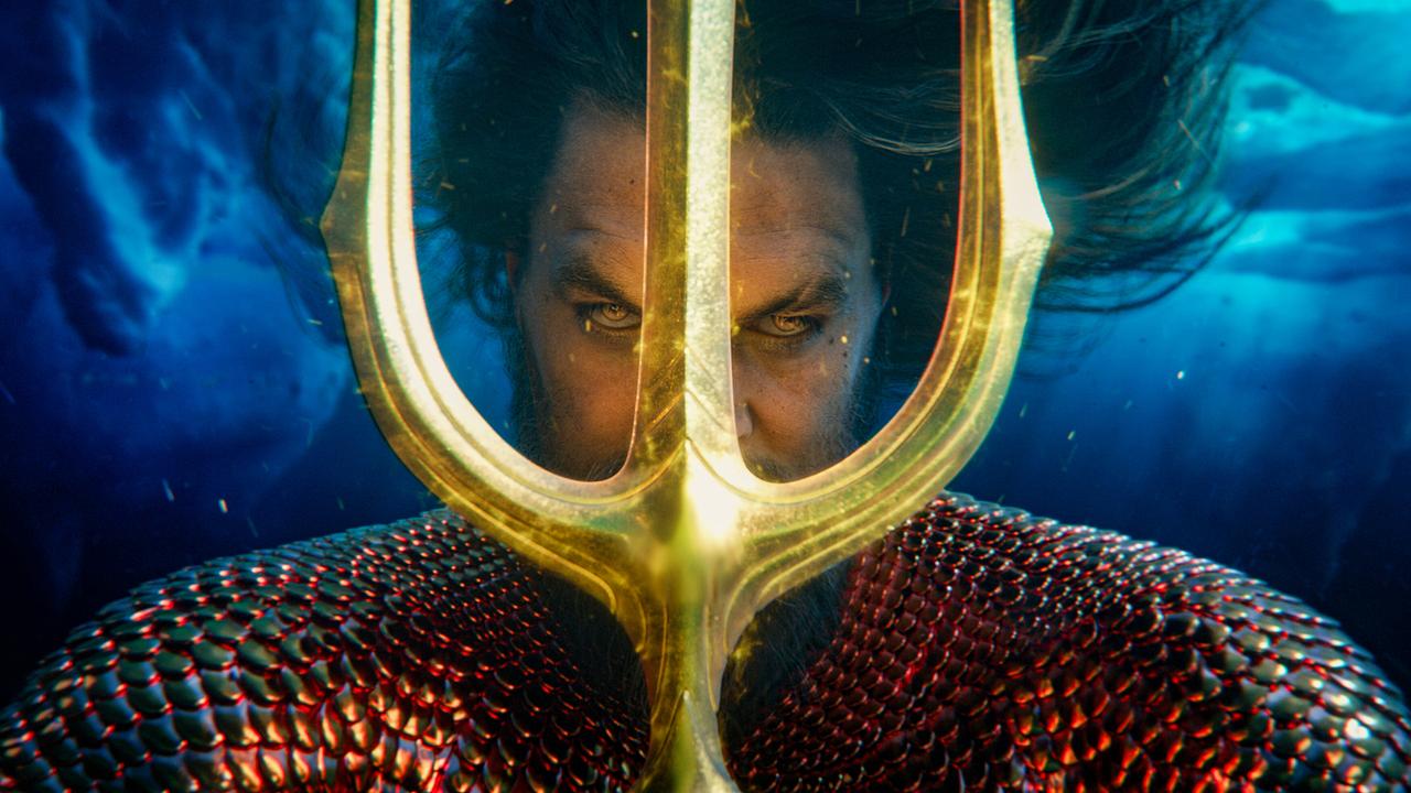Aquaman in schuppenartiger, rot schimmernder Rüstung hält einen goldenen Dreizack vor sein Gesicht, dahinter Unterwasserszenerie