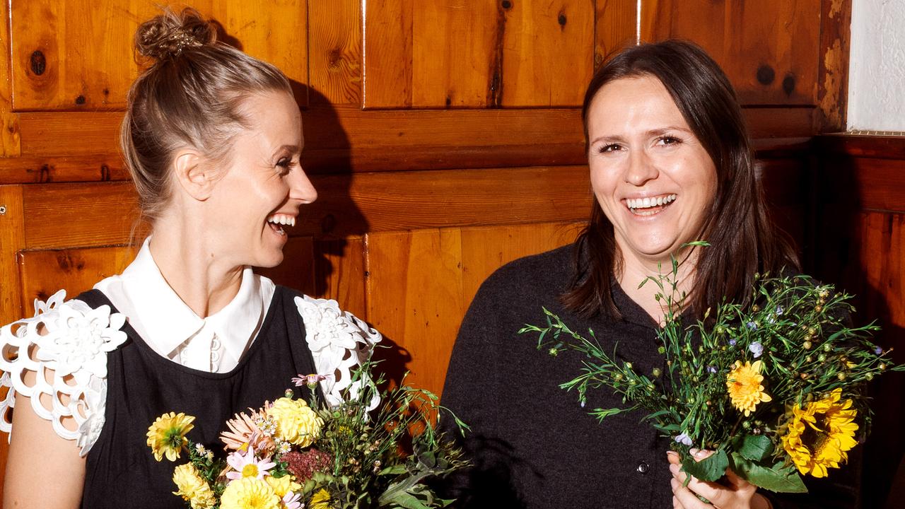 Kira Saskia Schinko mit Marie Rahofer sitzen im berühmten Linzer Leopoldistüberl an einem Holztisch und halten lachend selbstgebundene Blumensträuße.
