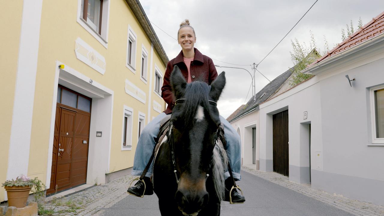 © ORF/Jenseide OG Kira Saskia Schinko reitet auf einem schwarzen Pferd durch eine schmale Dorfstraße zwischen Häusern.