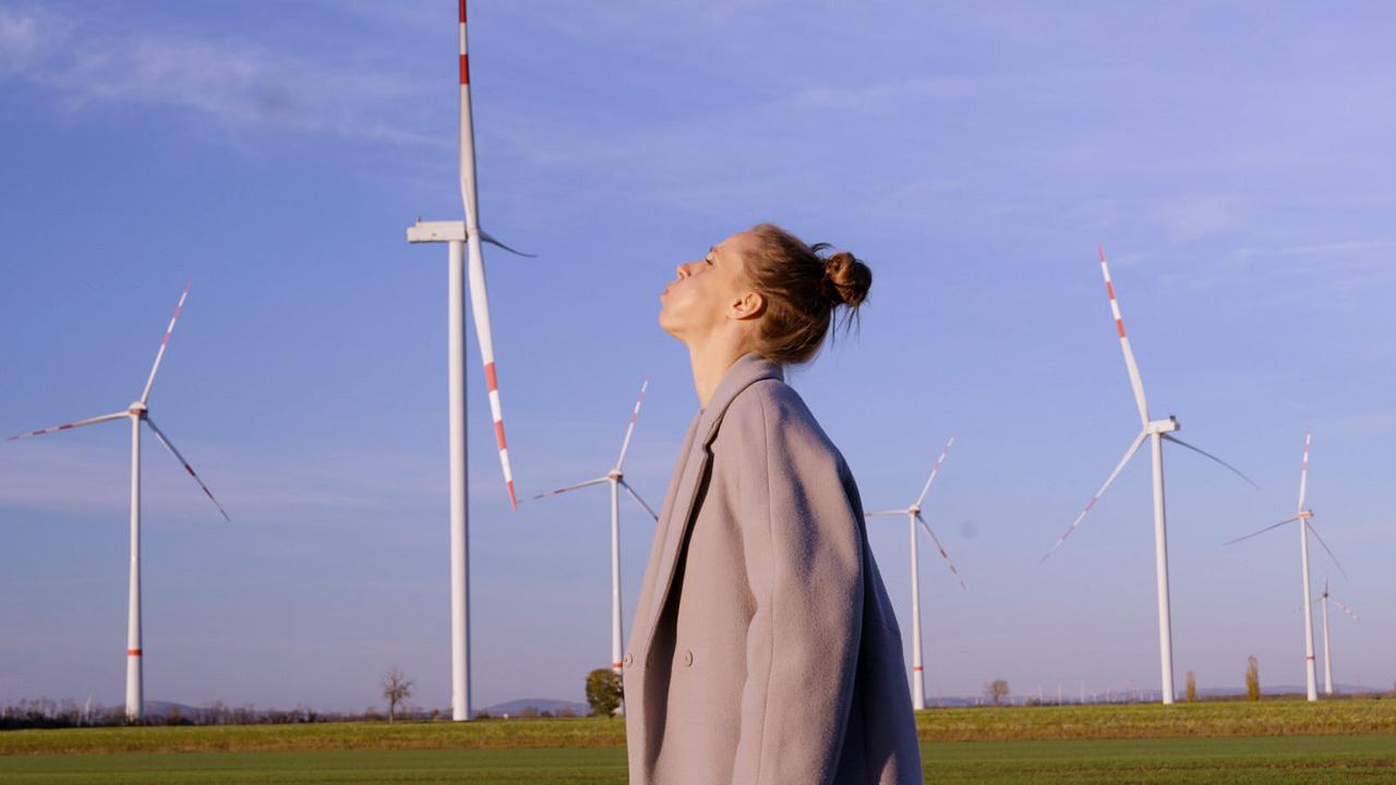 © ORF/Jenseide OG Kira Schinko steht vor einem Feld mit mehreren Windrädern unter blauem Himmel.