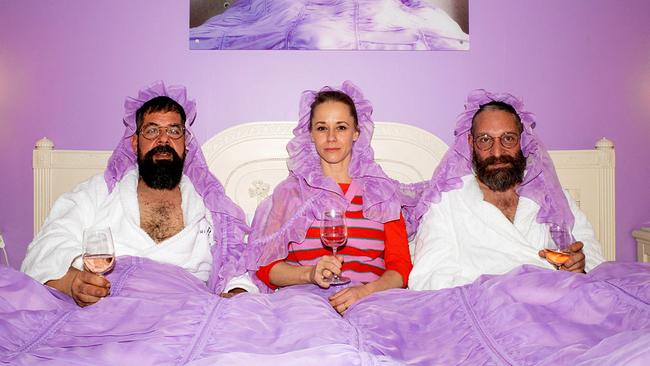 Kira Schinko im Pyjama sitzt mit Io Tondolo und Itshe Petz, beide in weißen Bademänteln, gemeinsam in einem Bett unter einer lilafarbenen Bettdecke und mit lilafarbenen Hauben am Kopf und halten ein Weinglas in der Hand.