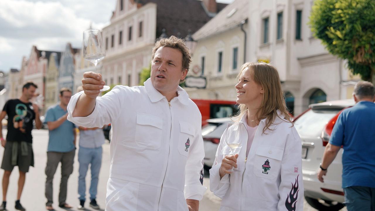 Kira Schinko mit Sternekoch Philipp Rachinger, beide in weißen Overalls, stoßen mit Weingläsern in einer belebten Altstadtstraße in Neufelden an, während im Hintergrund mehrere Passanten stehen.