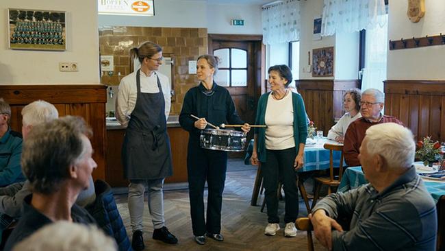 Im Gasthaus "Zur Palme" steht Kira Saskia Schinko mit einer kleinen Trommel zwischen mehreren sitzenden Gästen, während Theresia Palmetzhofer und eine weitere Frau danebenstehen.