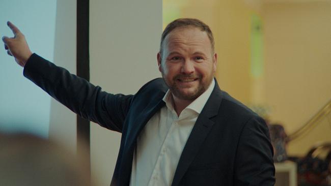 Ein Mann im dunklen Anzug mit weißem Hemd, dessen Kragen geöffnet ist, freundlich blickt er bei einer Präsentation zu den Zuhörenden, mit der rechten Hand zeigt er hinter sich auf den Bildschirm