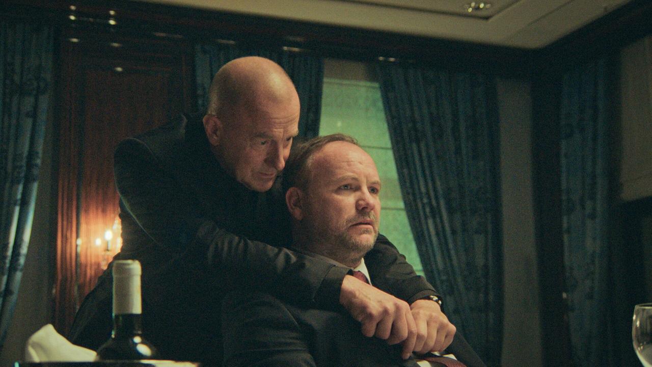 © ORF/UFA FICTION GmbH/Martin L. Ludwig Ein Mann im Anzug, Hemd und Krawatte sitzt beim Abendessen am Tisch, von hinten beugt sich ein Mann von hinten über ihn und flüstert eindringliche Worte ins Ohr