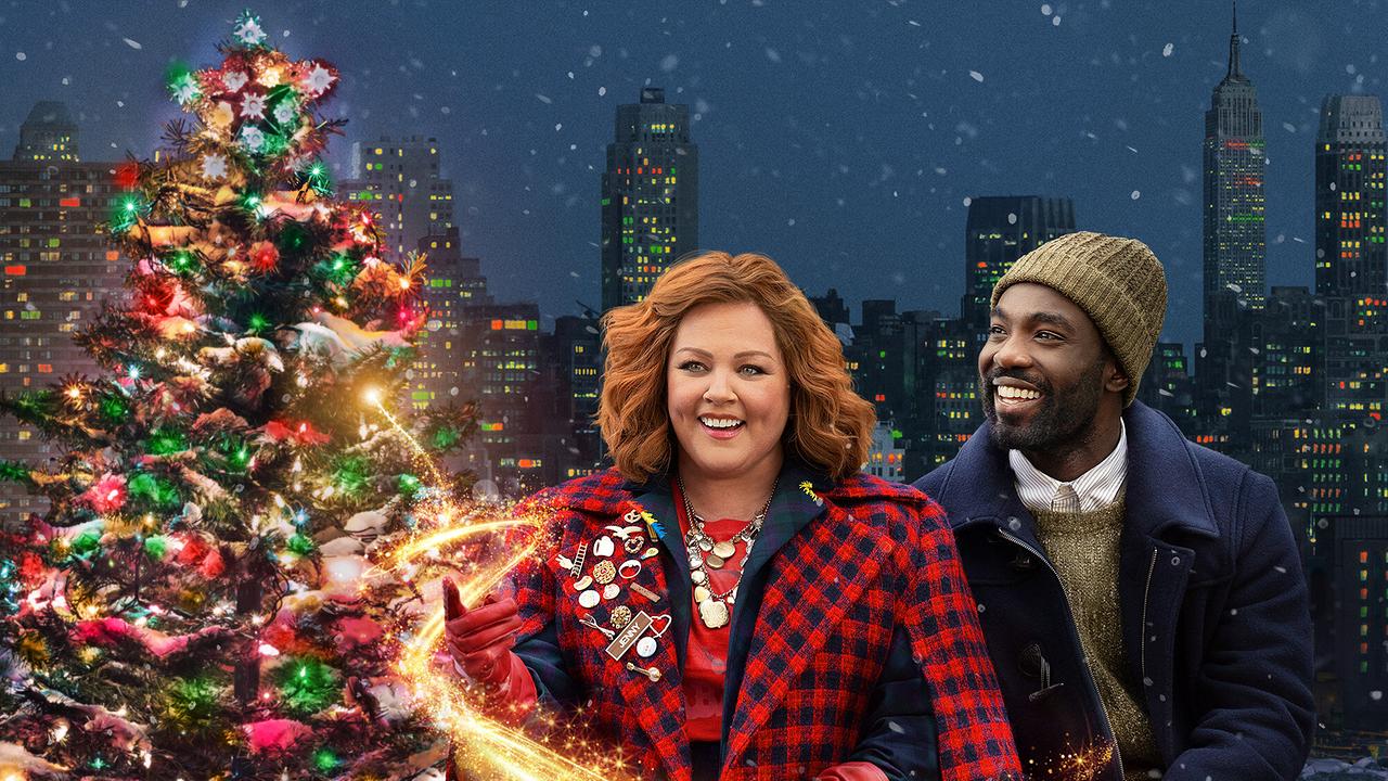 © ORF/Universal Pictures/Peacock Eine Frau im rot-schwarz-kariertem Mantel und ein Mann im blauen Mantel mit Haube stehen lächelnd neben einem festlich geschmückten Christbaum - vor nächtlicher Skyline einer Großsstadt