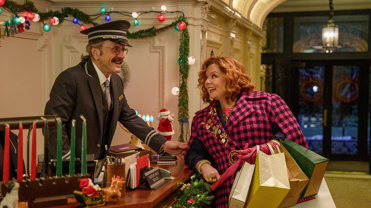 © ORF/Universal Pictures/Peacock/Stephanie Mei-Ling Eine Frau im pink-schwarz kariertem Mantel mit Einkaufstaschen im Arm spricht an einer festlich dekorierten Hotelrezeption mit einem uniformierten Angestellten