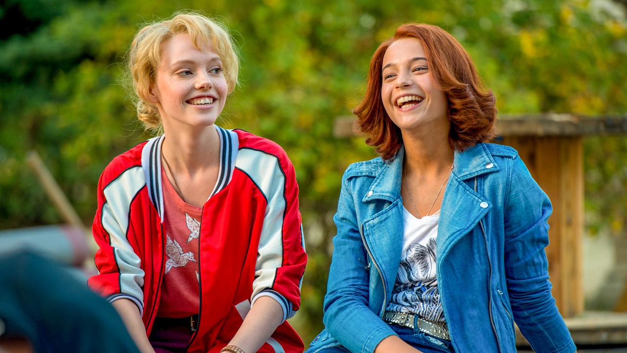 © ORF/DCM Film/Andreas Schlieter Lina Larissa Strahl als Bibi Blocksberg mit rotem T-Shirt und rot-weiß gestreifter Jacke und Lisa-Marie Koroll als Tina Martin in weißem T-Shirt mit blauer Jeans-Jacke, die beiden sitzen nebeneinander im Freien und lachen gelöst