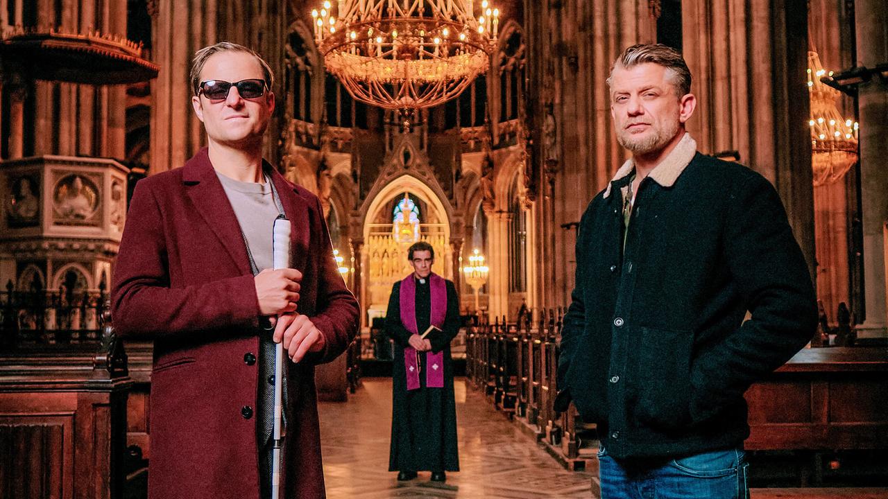Philipp Hochmair als Alexander Haller mit dunkler Brille und Blindenstock und Andreas Guenther als Nikolai Falk stehen in der Kirche, hinter ihnen im Gang zwischen den Bänken steht ein Pfarrer, im Hintergrund der Altar