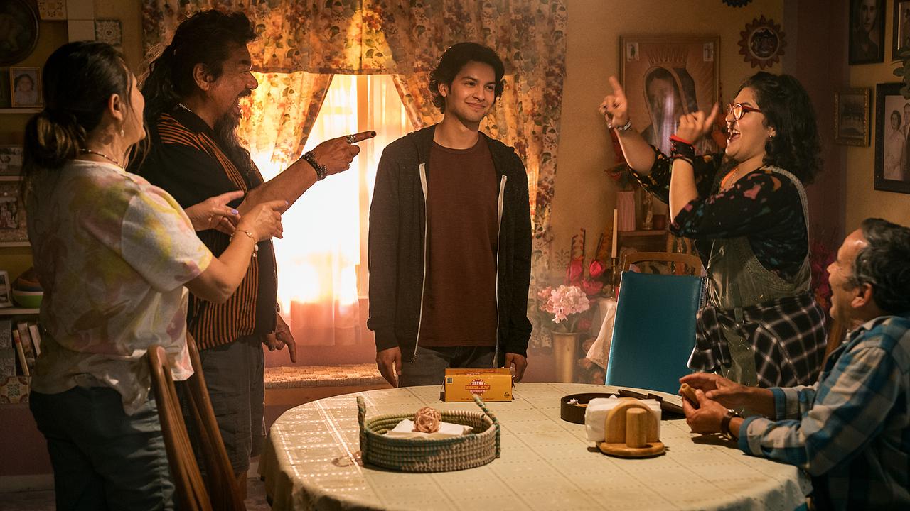 © ORF/Warner Bros. Pictures Xolo Maridueña steht als Jaime Reyes lächelnd in einem Wohnzimmer, zwei ältere Paare stehen um den Wohnzimmertisch und deuten fröhlich auf ihn und feiern ihn