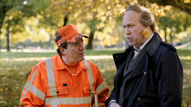 Robert Palfrader als Straßenkehrer im orangenen Arbeitsanzug und Nicholas Ofczarek im dunklen Mantel und Anzug stehen in einem herbstlichen Park