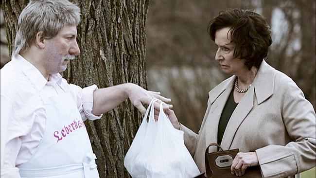 Robert Palfrader in weißer Kleidung und einer Schürtze mit der Aufschrift "Leberkäspepi" überreicht Barbara Petritsch ein Plasticksackerl vor einem Baum in einem Park.
