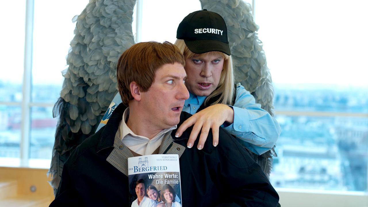 Vor einer Glasfront in einem hellen Raum umarmt Nicholas Ofczarek, mit großen Engelsflügeln und Security‑Kappe am Kopf, von hinten Robert Palfrader, der ein Magazin mit dem Titel „Wahre Werte: Die Familie“ in der Hand hält.