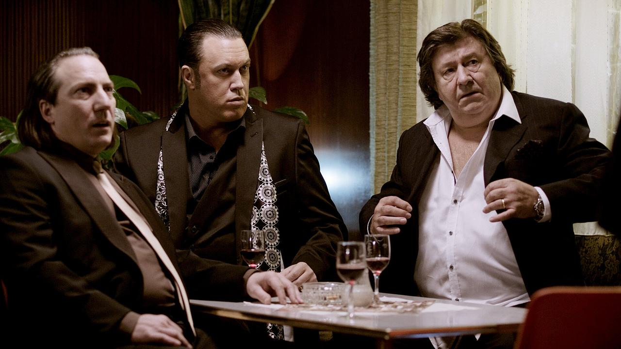 © ORF/Superfilm Robert Palfrader und Nicholas Ofczarek, beide im schwarzen Anzug und dunklem Hemd, sitzen mit Hanno Pöschl, ebenfalls im Anzug und weiße, Hemd, an einem Tisch in einem Lokal, vor ihnen stehen Weingläser,.