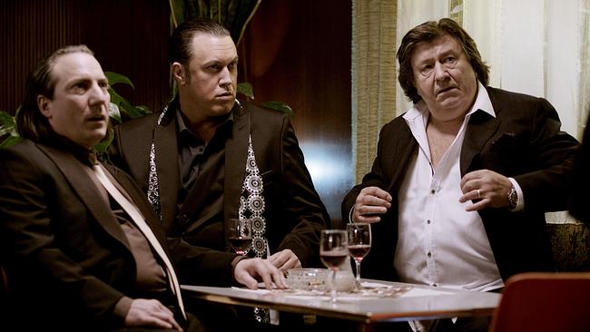 Robert Palfrader und Nicholas Ofczarek, beide im schwarzen Anzug und dunklem Hemd, sitzen mit Hanno Pöschl, ebenfalls im Anzug und weiße, Hemd, an einem Tisch in einem Lokal, vor ihnen stehen Weingläser,.