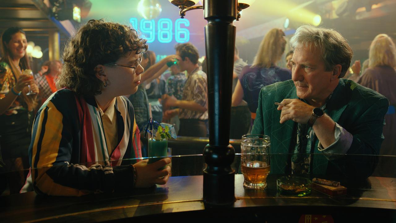 B.B. und Gerri Tschach sitzen mit einem Getränk an einer Bar und unterhalten sich in einem lebhaften Nachtclub, in dem an der Wand bei der Tanzfläche die Zahl 1986 projiziert wird.
