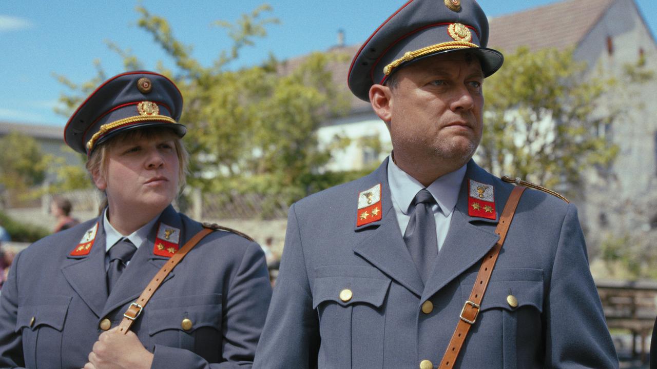 Polizistin Gerti und Polizist Hannes stehen bei Sonnenschein im Freien in einem Dorf und schauen nach rechts.