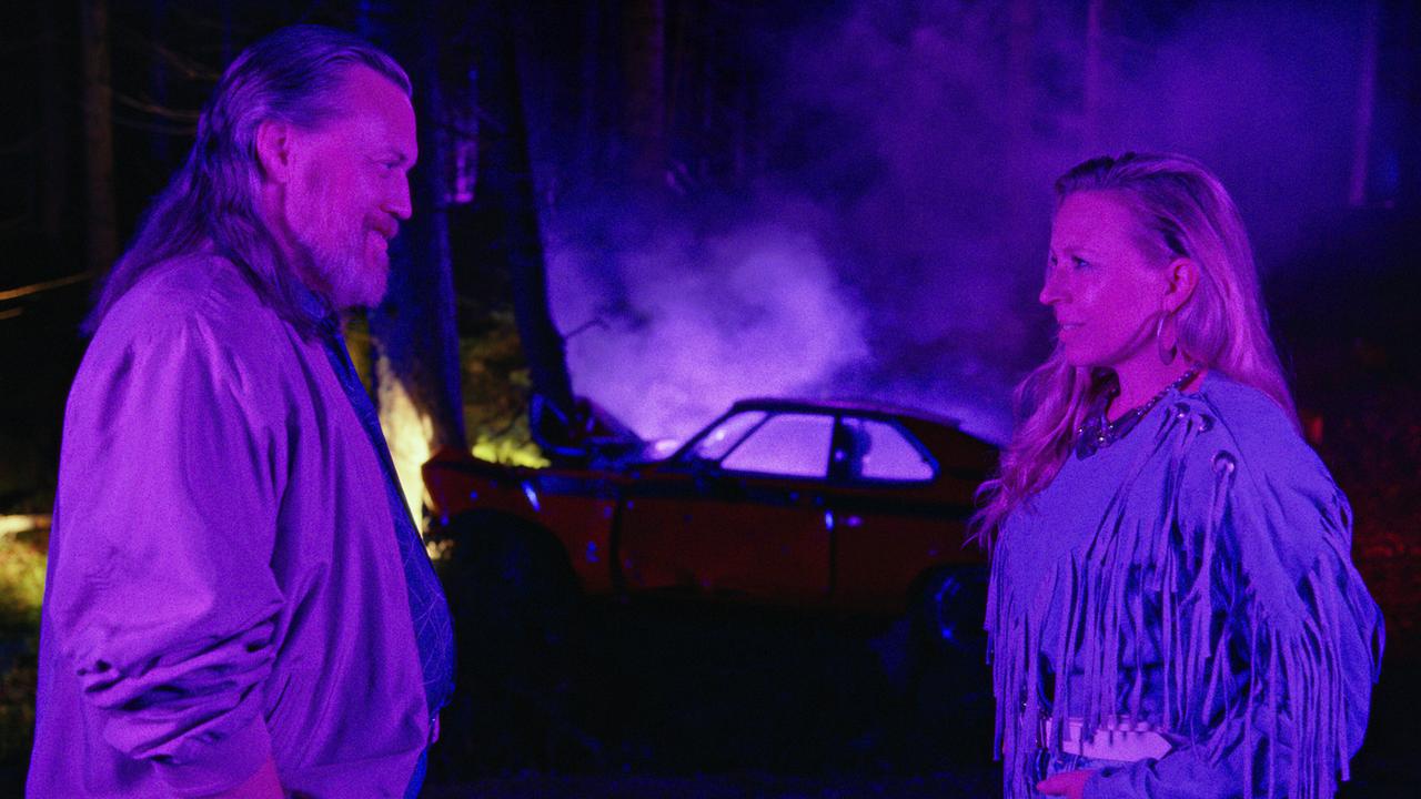 Richard Pfeisinger und Elfi Pfeisinger stehen sich lächelnd nachts im Wald gegenüber, im Hintergrund ein zerstörtes Auto und Rauch.