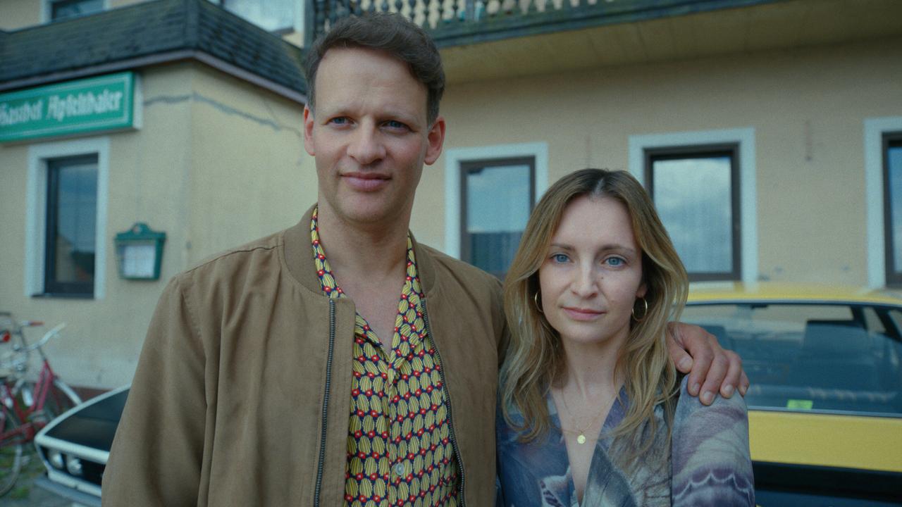 Kevin hat einen Arm um Babs Tschachs Schulter, während sie bei Tag vor einem Gasthaus stehen, hinter ihnen ist ein parkendes Auto zu sehen.