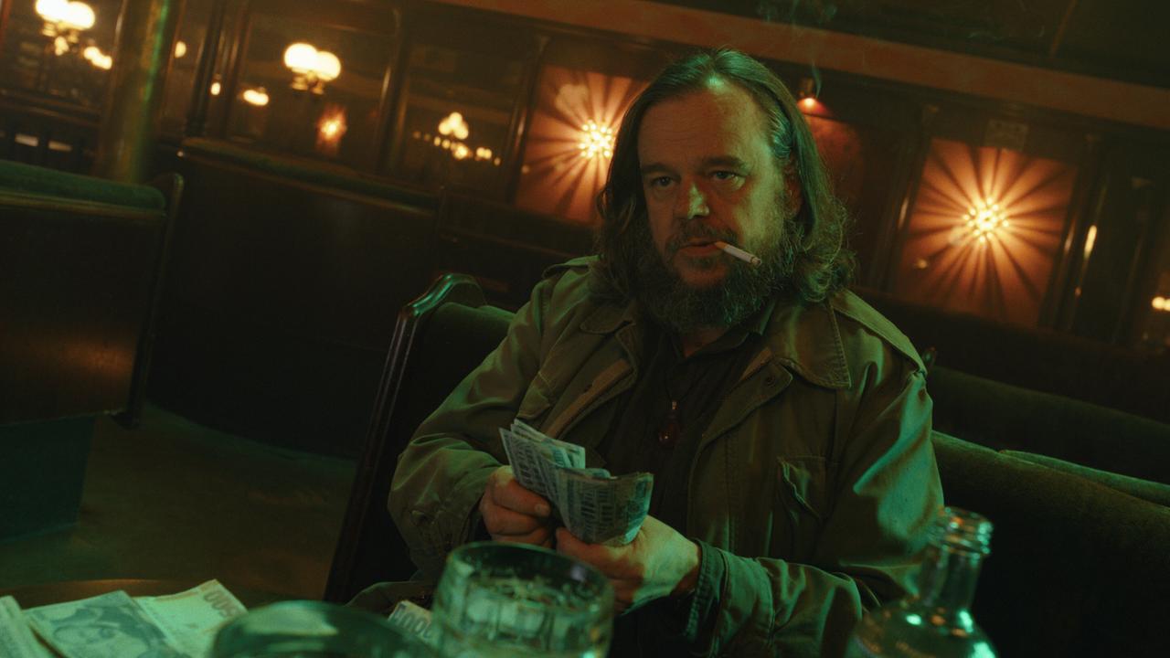 Reinhard Matussek sitzt allein, mit einer Zigarette im Mund, in einem dunklen Club und zählt Geldscheine auf einem Tisch.