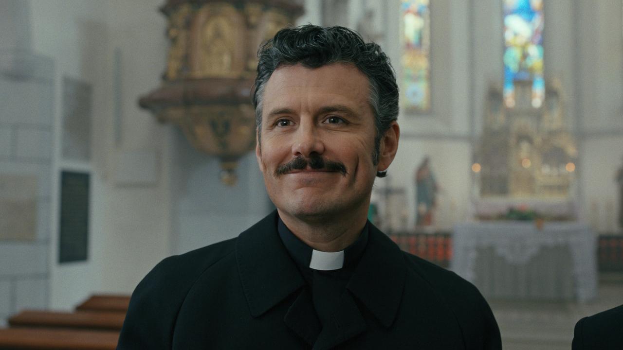 Banyardi in geistlicher Kleidung steht lächelnd im Inneren einer Kirche.