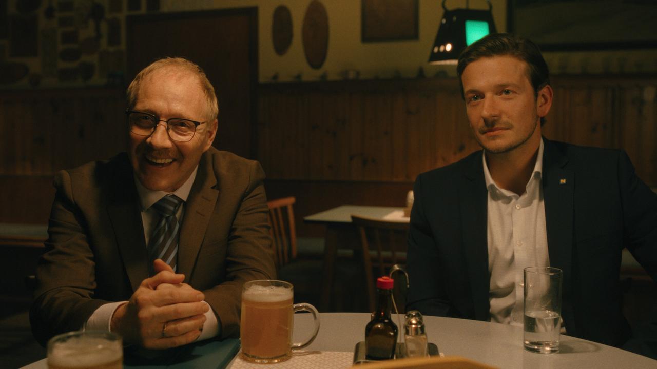 Katzlbrunner und Sigmund sitzen nebeneinander in einem Wirtshaus bei einem Bier bzw. einem Glas Wasser.