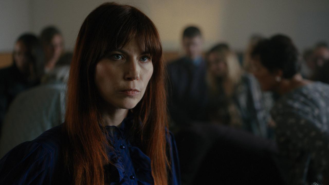 Deborah Feist steht in einem dünkleren Raum, im Hintergrund sitzen unscharf mehrere Menschen in Reihen.