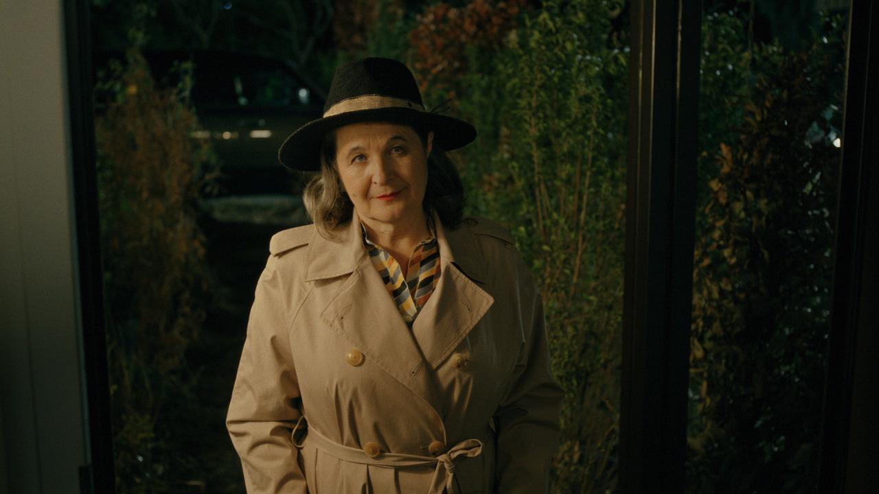 Herta Tschach im Trenchcoat und Hut steht nachts vor einer Glasfront mit Pflanzen dahinter.