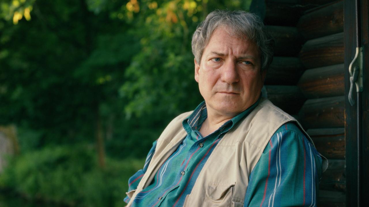 © ORF/Superfilm Gerri Tschach sitzt vor einer Holzhütte im Grünen.