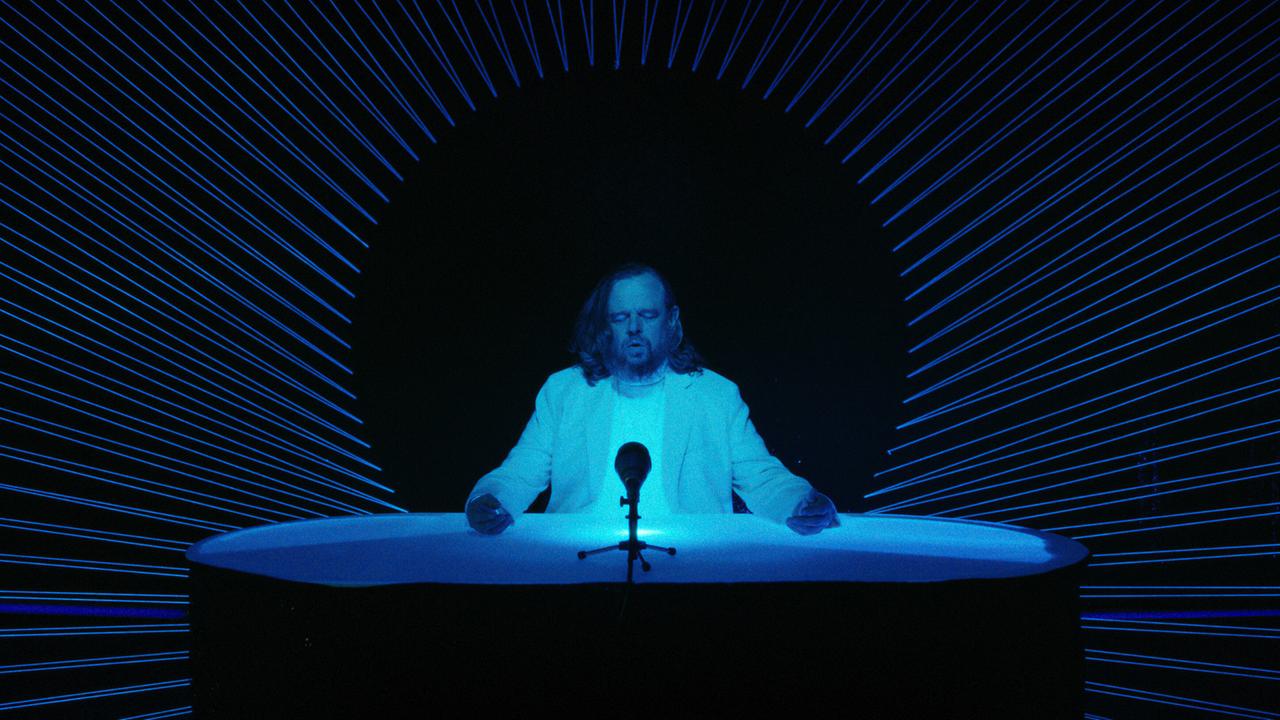 © ORF/Superfilm Reinhard Matussek sitzt an einem futuristisch beleuchteten Tisch mit Mikrofon in einer dunklen, blau inszenierten Studioszene.