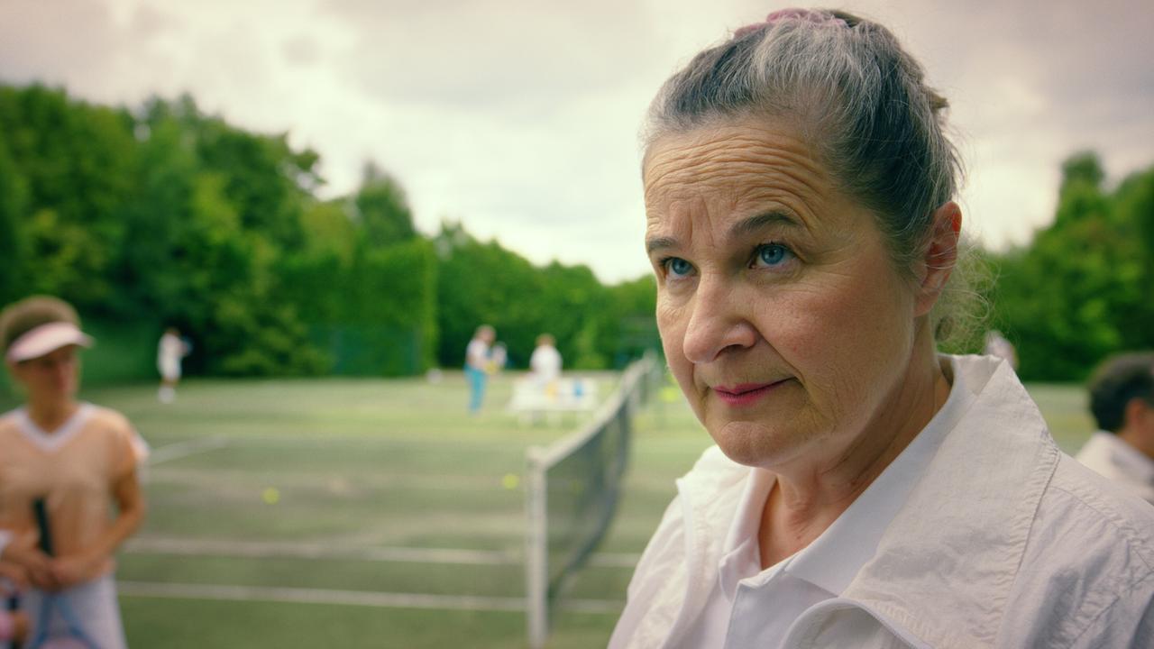 © ORF/Superfilm Herta Tschach im Vordergrund steht vor Tennisplätzen im Grünen, auf dem Meschen spielen.