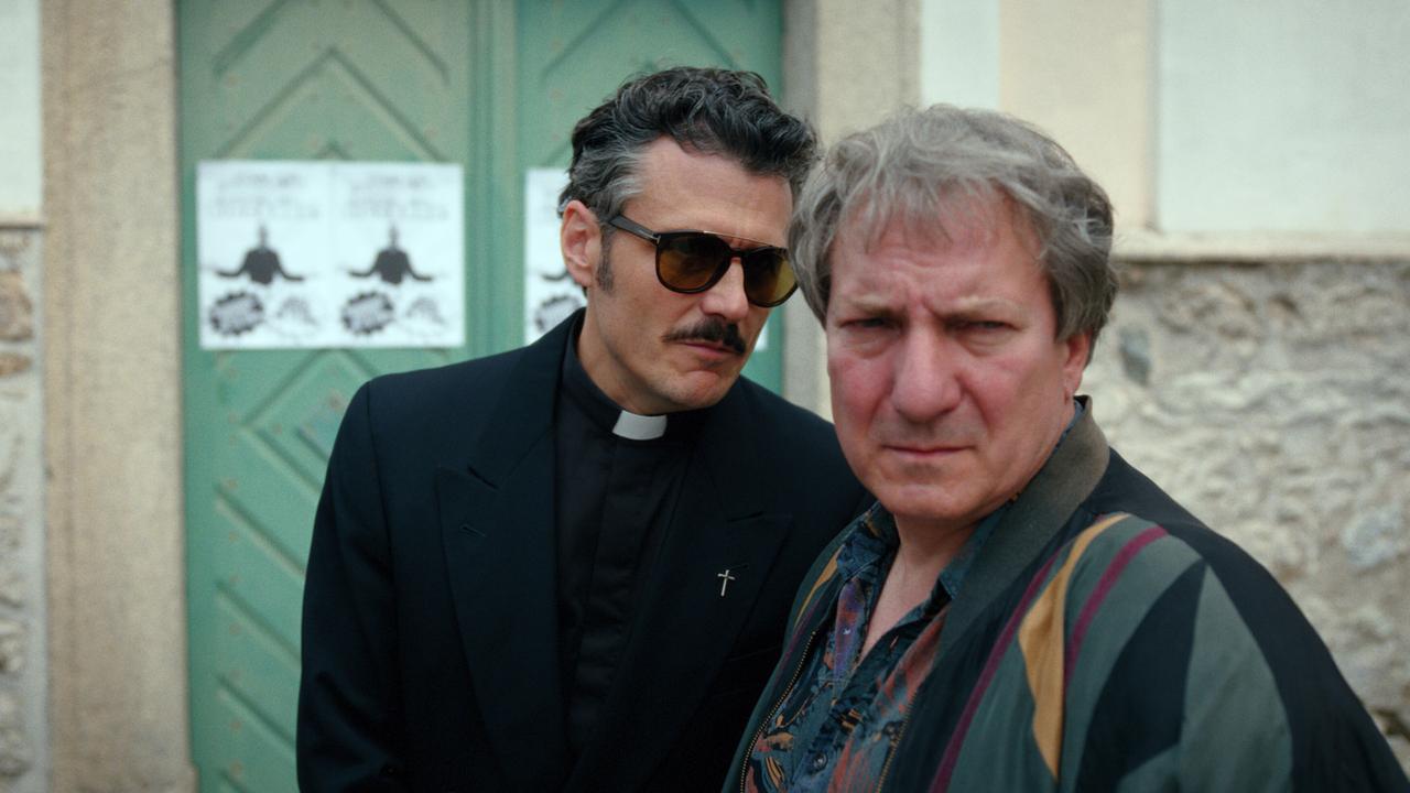 © ORF/Superfilm Priester Banyardi mit Sonnenbrille und Gerri Tschach stehen nebeneinander vor dem Eingang eines kirchlichen Gebäudes.