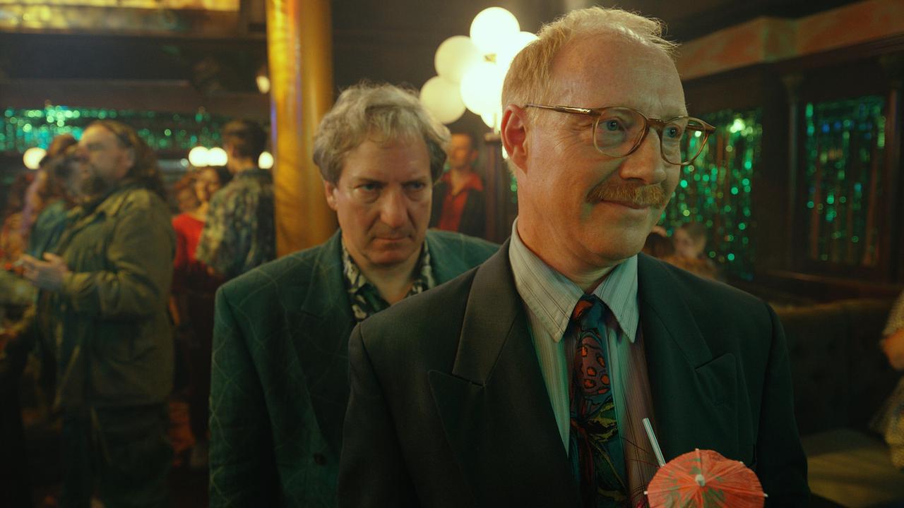 © ORF/Superfilm Gerri Tschach und Katzlbrunner in 80er-Anzügen stehen in einem Tanzlokal mit Cocktails, während im Hintergrund gefeiert wird.