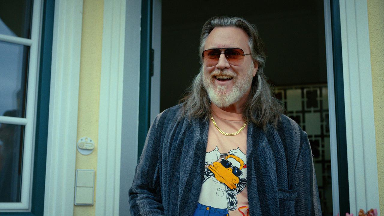 © ORF/Superfilm Richard Pfeisinger mit langem Haar und Vollbart steht im Türrahmen eines Hauses und trägt einen Morgenmantel über einem T‑Shirt.