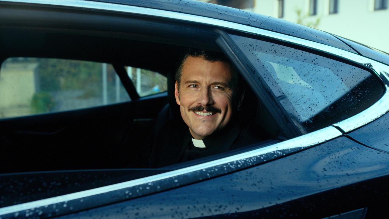 © ORF/Superfilm Priester Banyardi sitzt bei Regen auf dem Rücksitz eines dunklen Autos und blickt lächelnd aus dem Fenster.