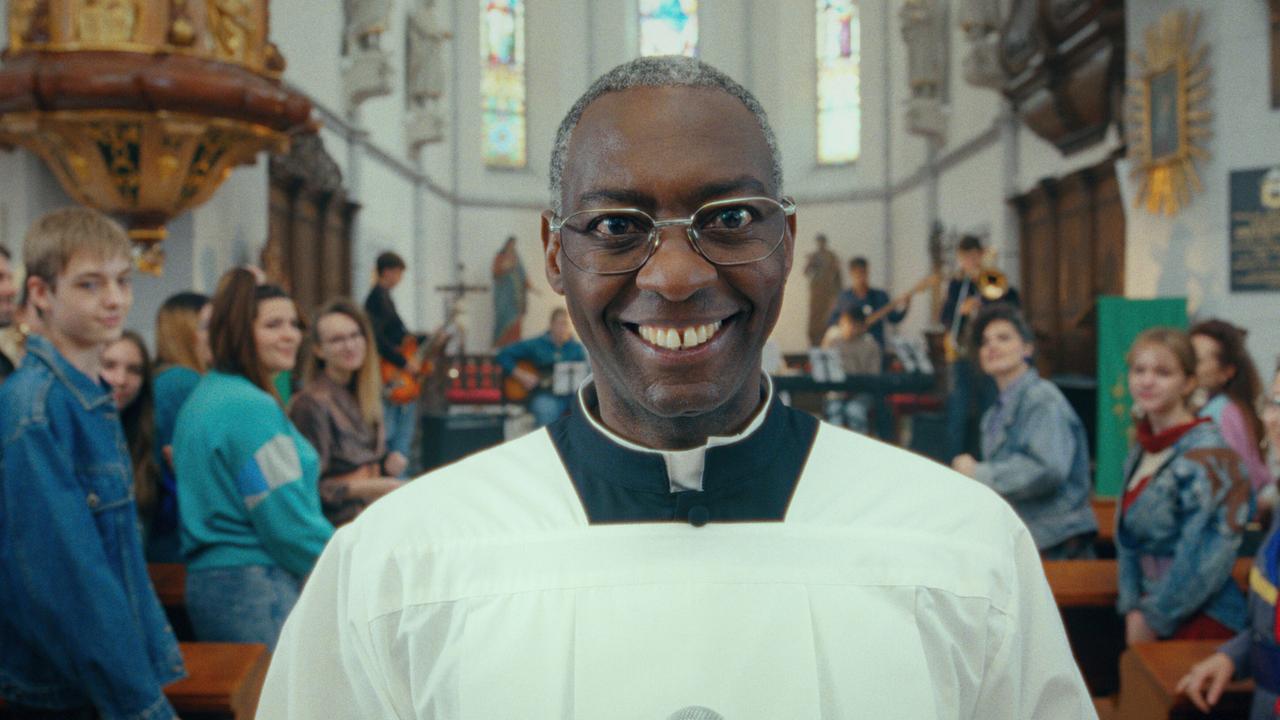 © ORF/Superfilm Priester Riccardo steht lächelnd in der Kirche im Vordergrund, während im Hintergrund eine Band vor der Gemeinde spielt.