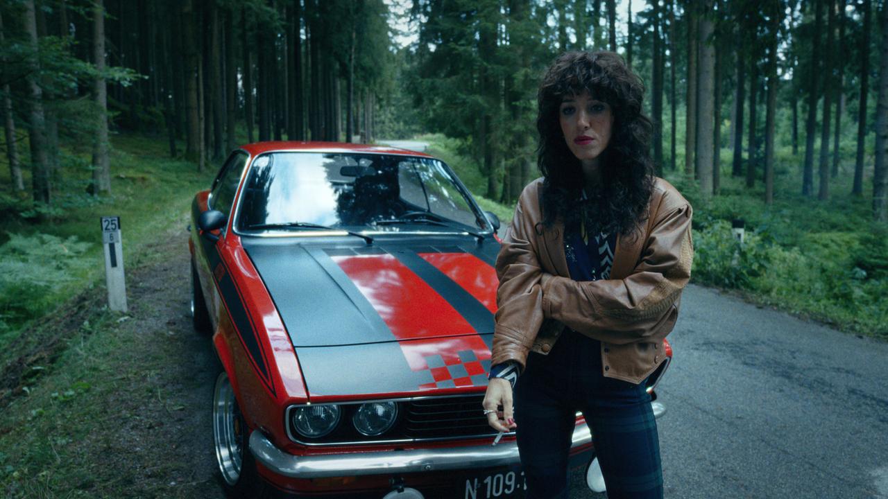 © ORF/Superfilm Ronda steht vor einem roten Oldtimer auf einer schmalen Waldstraße und hält eine Zigarette in der Hand.