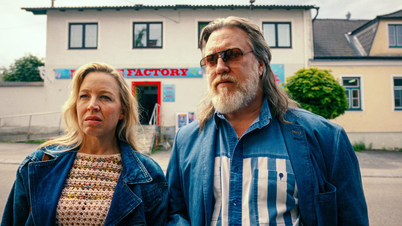 Elfi Pfeisinger und Richard Pfeisinger stehen tagsüber nebeneinander vor einer Häuserzeile mit buntem Schriftzug und blicken ernst nach vorne.