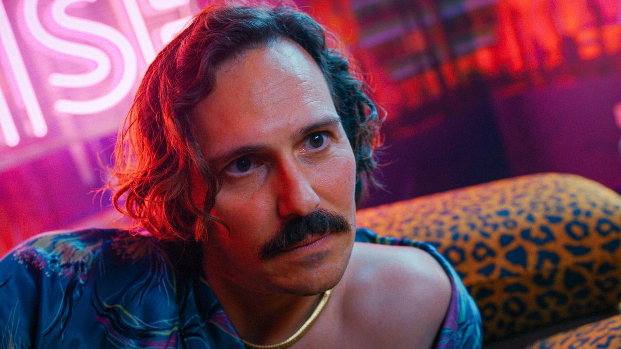 Ronnie  sitzt in einem neonbeleuchteten Raum mit pink‑roter Lichtstimmung und lehnt sich entspannt auf ein Sofa mit Leopardenmuster.