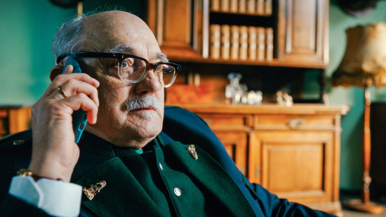 Kortner im Trachtenanzug  sitzt in einem Wohnzimmer und telefoniert am Handy vor einem hölzernen Regal mit Büchern und Dekoration.